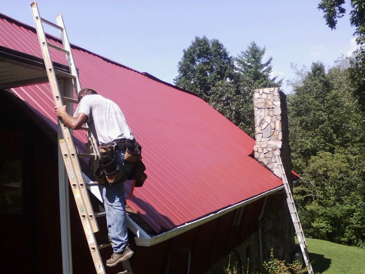 Roofer on a ladder beside a red metal roof and stone chimney — Soffit & Fascia Repair by Roofing Brothers in Dow City